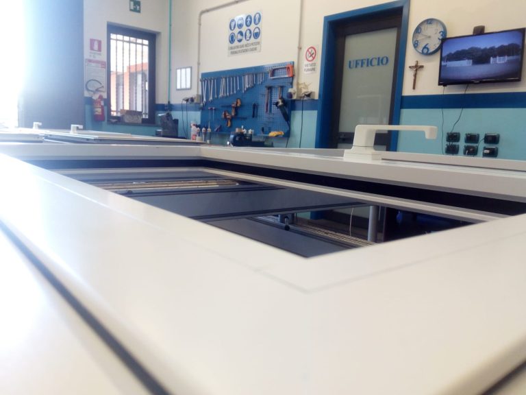Interno di un'officina o laboratorio specializzato nella produzione di infissi in alluminio, dove si notano diverse finestre in fase di assemblaggio su un banco da lavoro